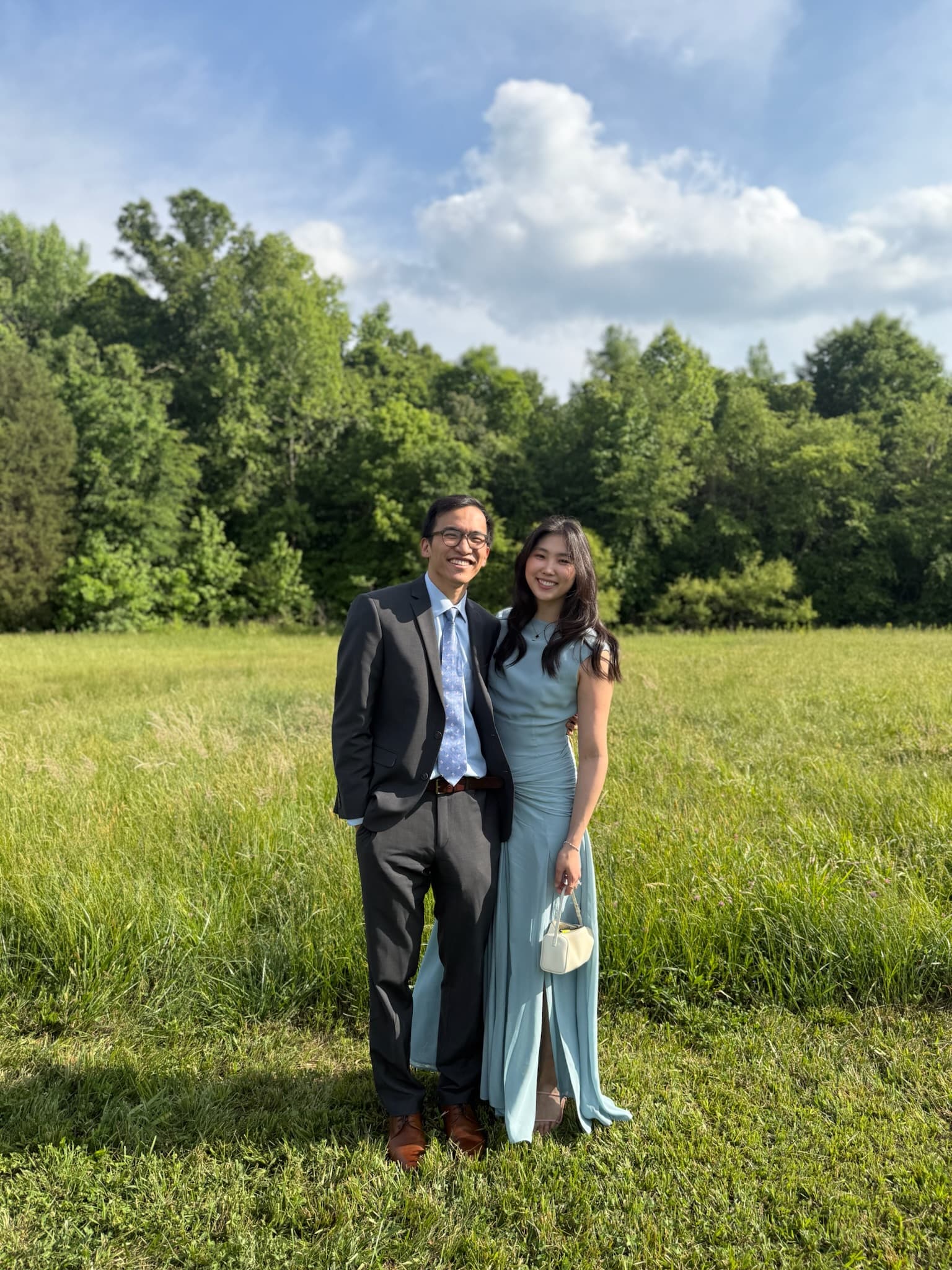 Dressed for a wedding, standing in a summer field against a tree line.