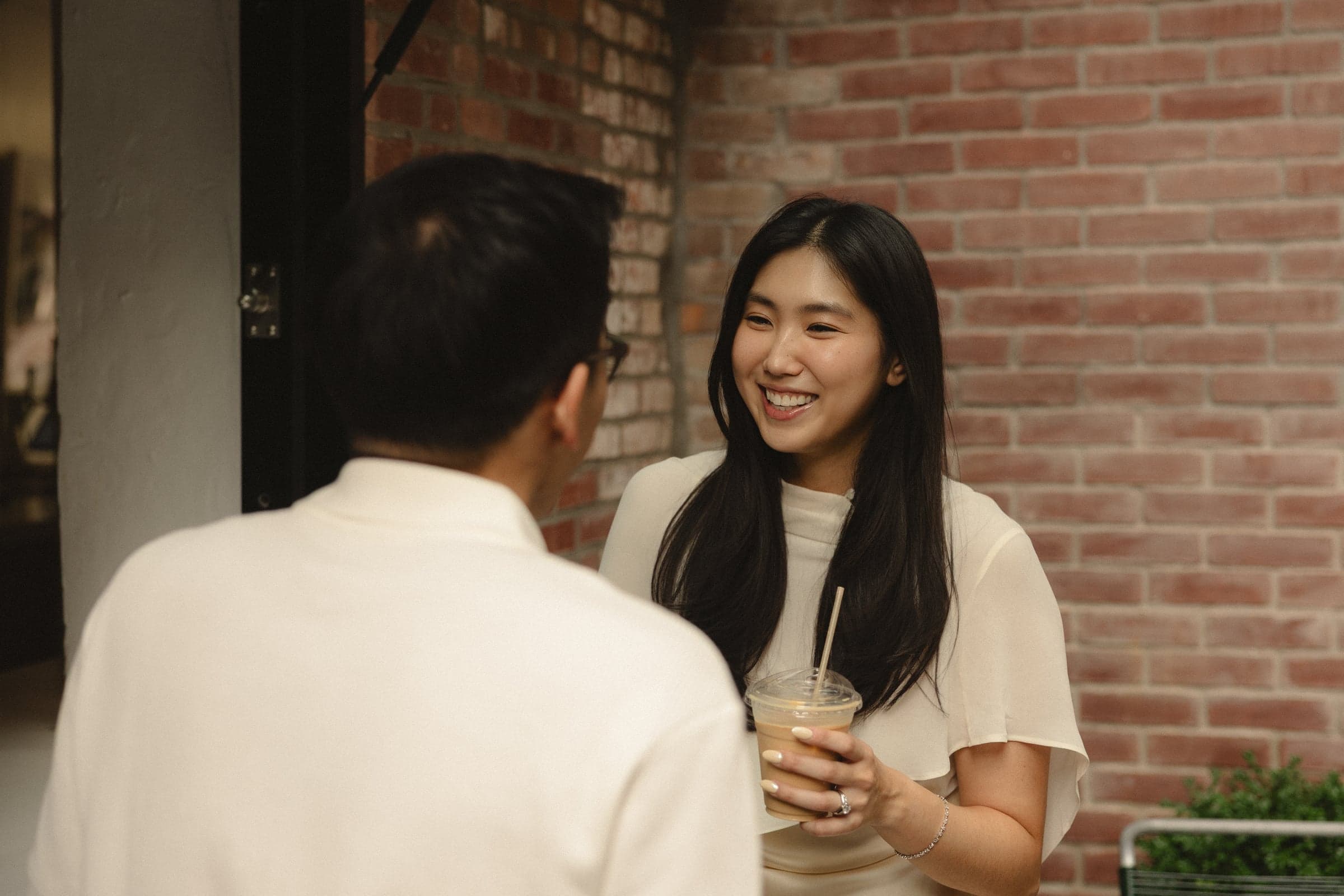 Laughing with an iced coffee in hand, against a brick wall.