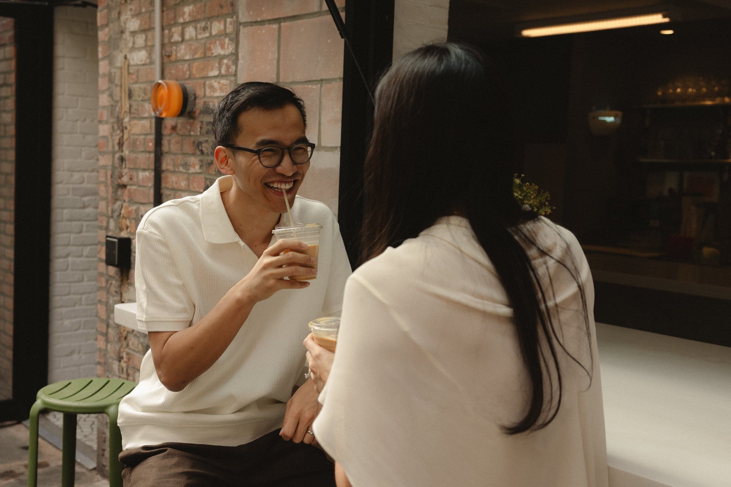 Iced coffees at a cafe table, mid-laugh against the brick.