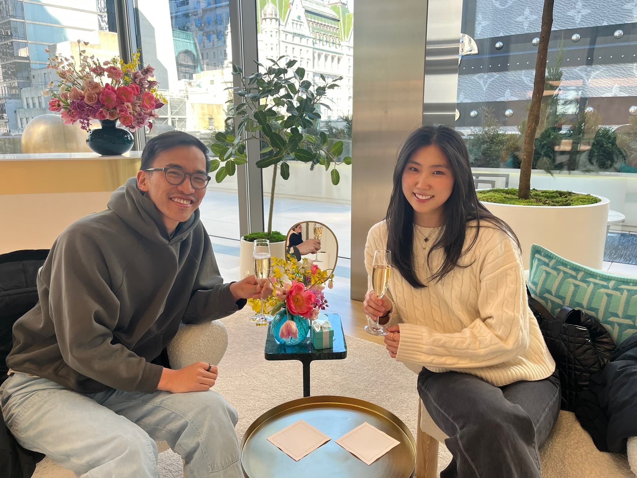 Raising champagne in a bright lounge, a table of flowers between us.