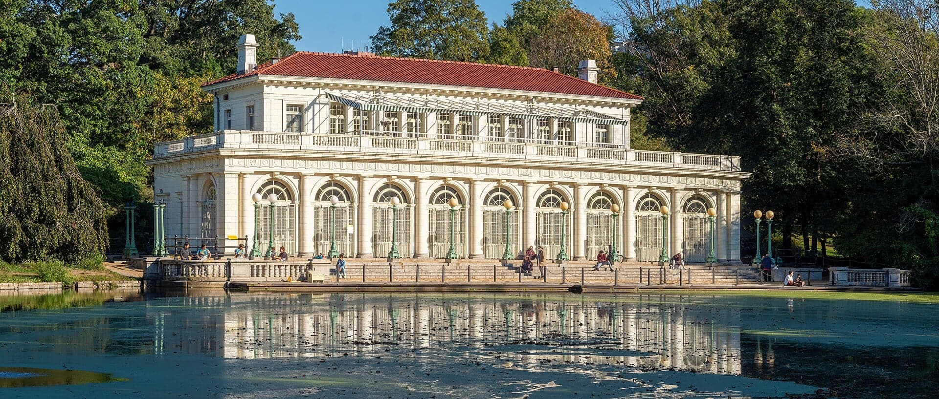 The Prospect Park Boathouse across the water: a white pavilion with an arcade of tall arched windows under a red tile roof, mirrored in the lake.