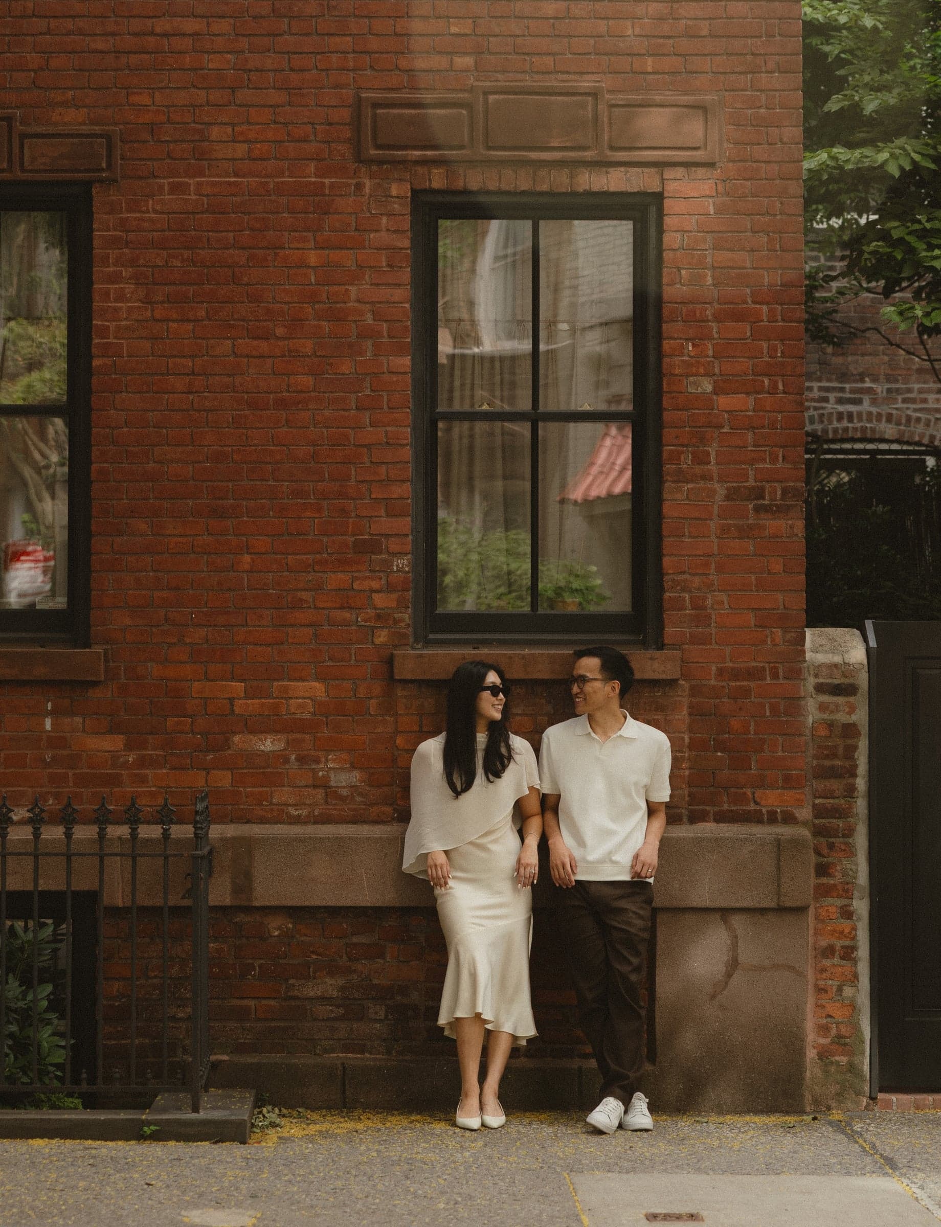 Leaning against a brick wall under a tall window, looking at each other.